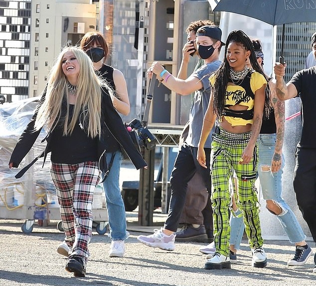 À esquerda Avril Lavigne com casaco preto e calça quadriculada avermelhada e à direita Willow com blusa amarela e preta e calça quadriculada nas mesmas cores. Elas andam descontraídas e sorridentes pelo set de gravação com pessoas atrás.