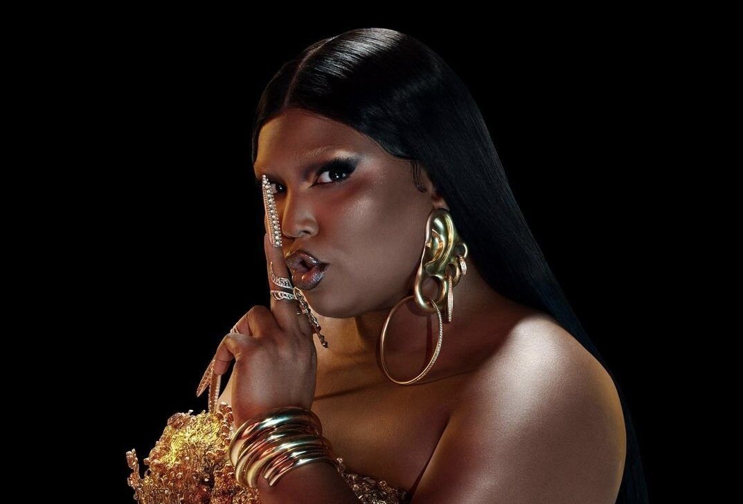 "Rumors": Lizzo divulga lançamento para este mês em capa com fundo preto, aparecendo com acessórios dourados em sinal que pede silêncio.